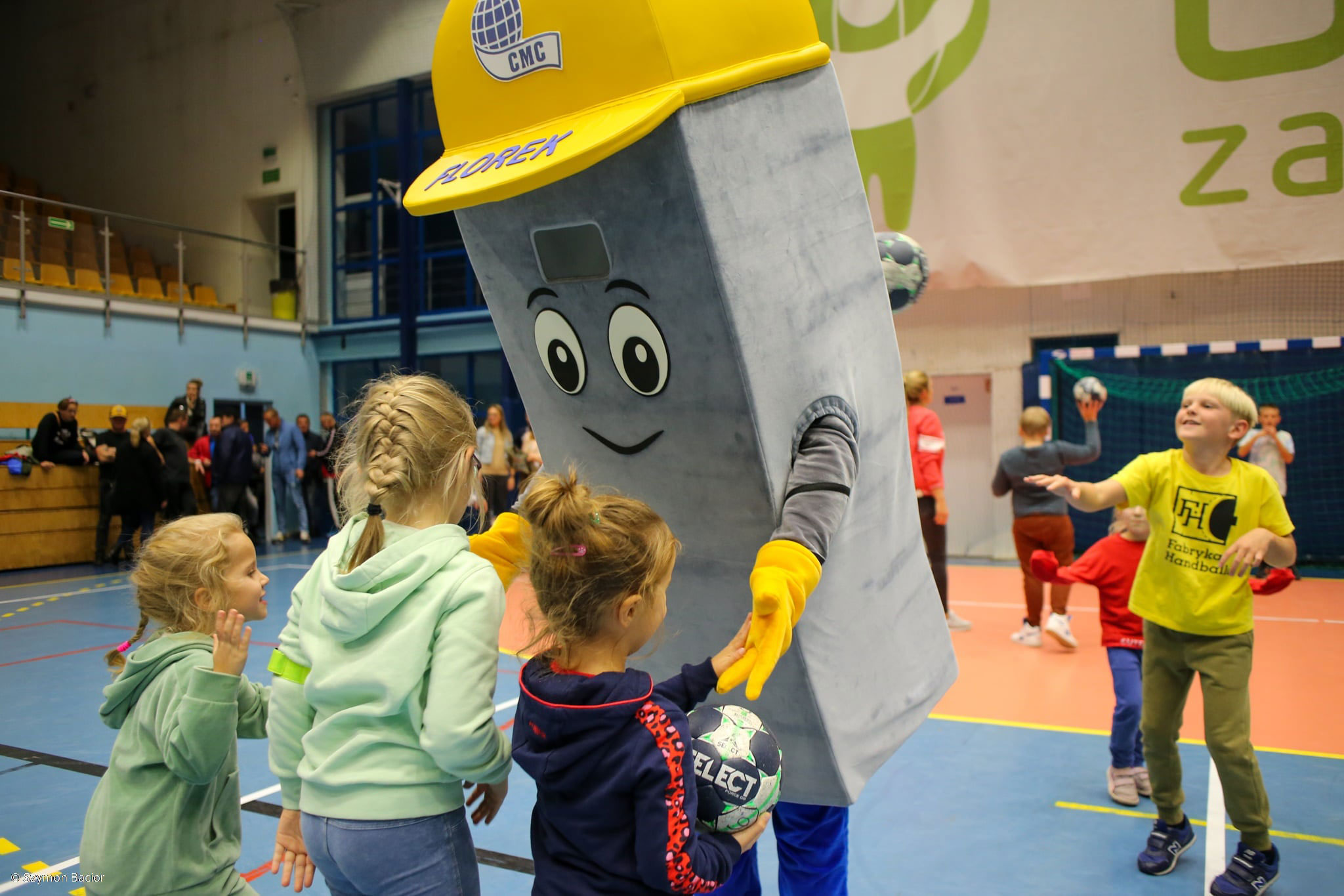 Kostium reklamowy w postaci hutnika pochylający nie trójką dzieci. Na drugim palnie inne osoby, w tle hala sportowa